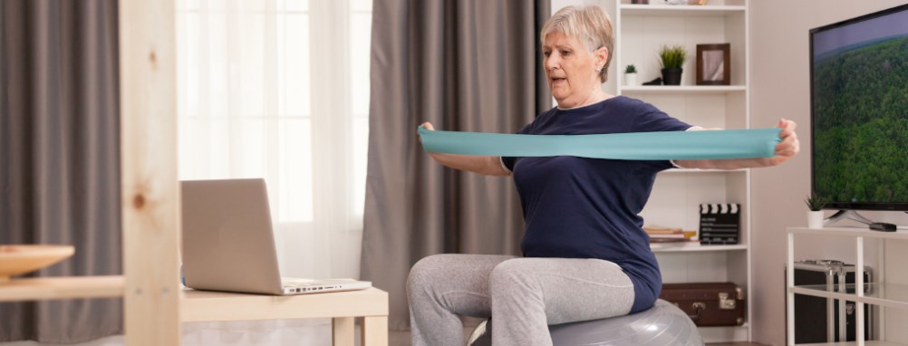 Older adult woman on exercise ball with resistance band following virtual class on laptop